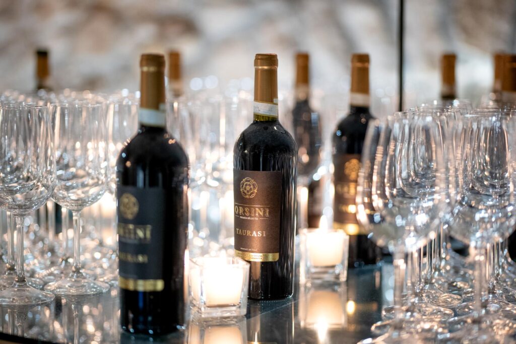 Bottiglie di vino Taurasi Orsini su tavolo riflettente, circondate da calici di cristallo e candele accese, in un'elegante atmosfera da degustazione.